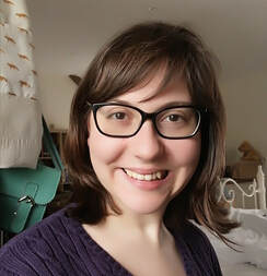 Amy is smiling into the camera, she has brown hair with a fringe and is wearing glasses.